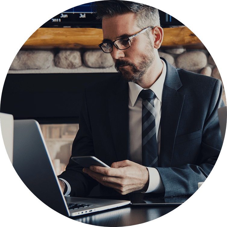 A man in a suit looks at his phone while sitting in front of a laptop